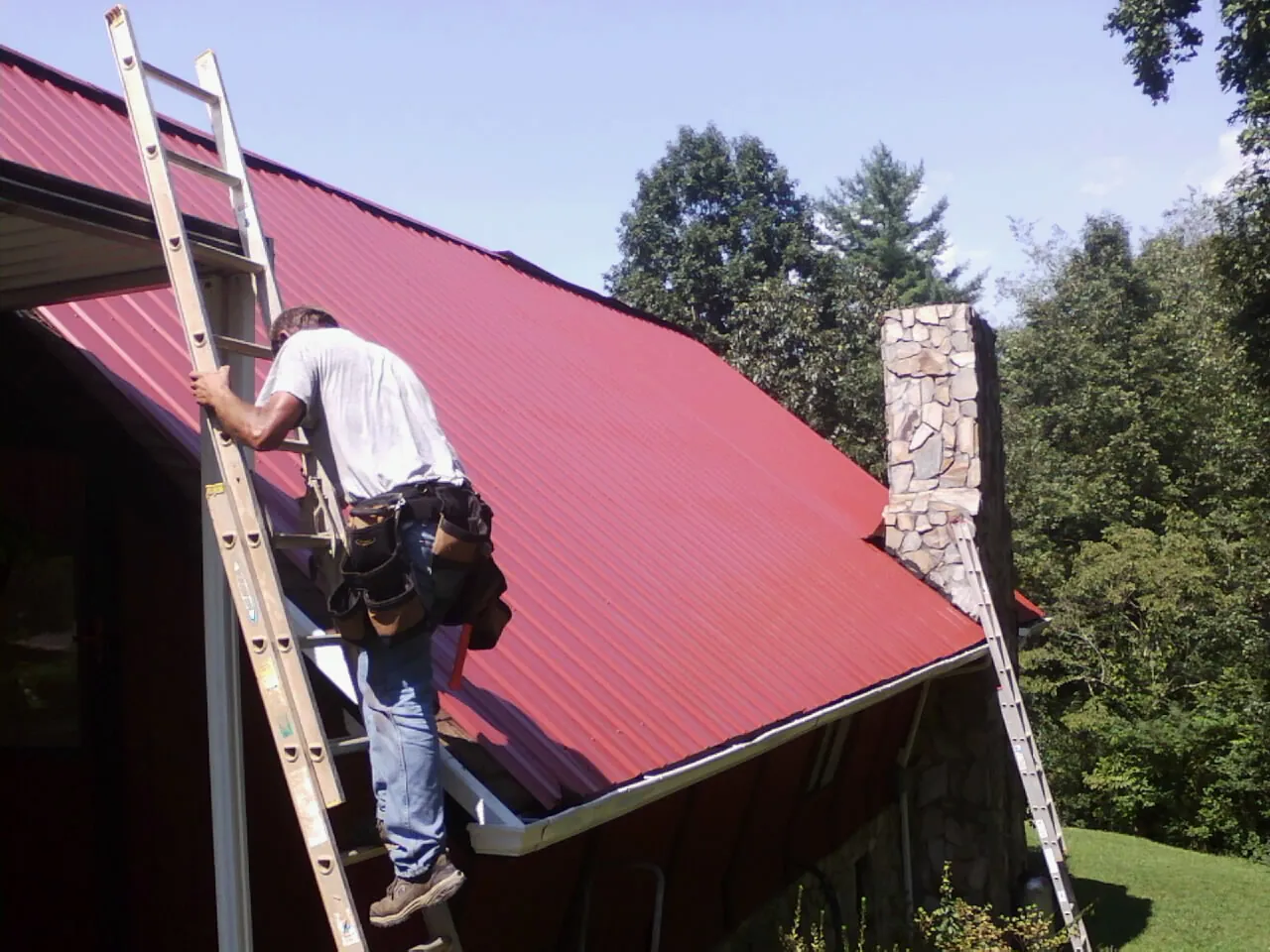 Roofer on a ladder beside a red metal roof and stone chimney — Soffit & Fascia Repair by Roofing Brothers in Gales Ferry